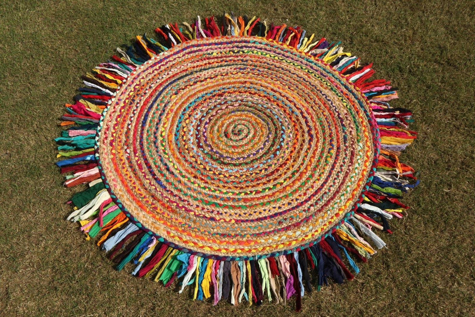 A round braided rag rug made of rainbow fabric strips with a long multicoloured fringe, laid flat on grass