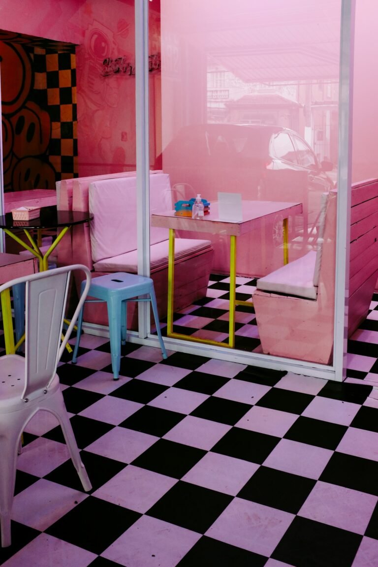 A pink-walled café with a pink and black checkerboard floor, a pale blue metal stool, a white metal chair and a table on yellow legs