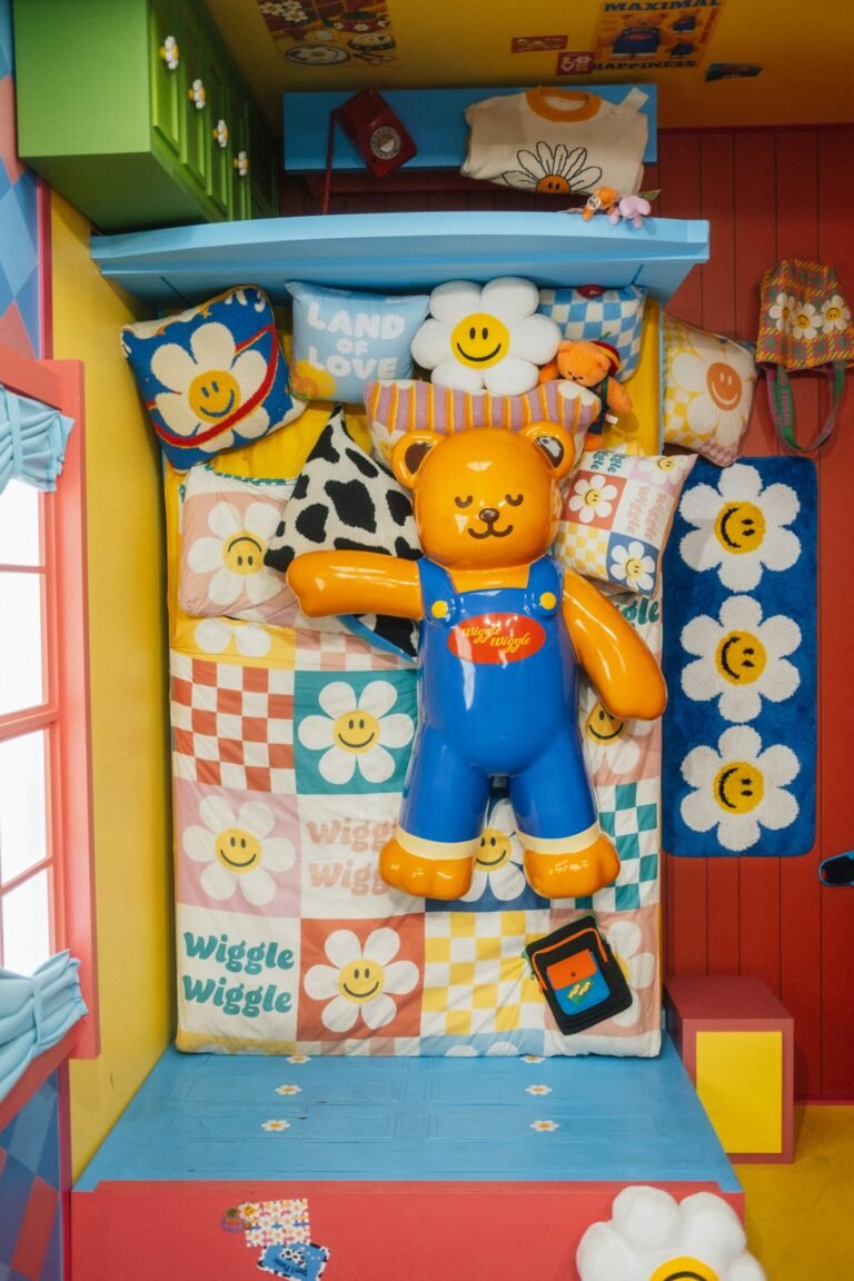 Overhead view of a bed piled with patchwork bedding of smiling daisies and checkerboard squares, a cow-print cushion and a giant orange bear figure in blue overalls, between red and yellow walls and a blue daisy rug