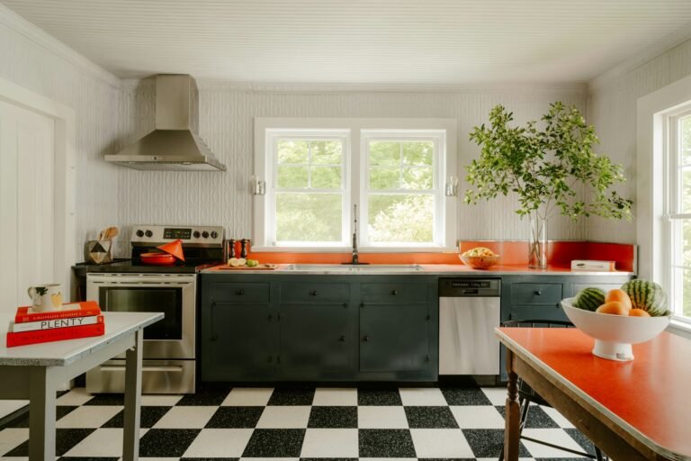 A kitchen with dark green cabinets, an orange worktop and splashback, a black-and-white checkerboard floor and a red-topped table holding a bowl of fruit