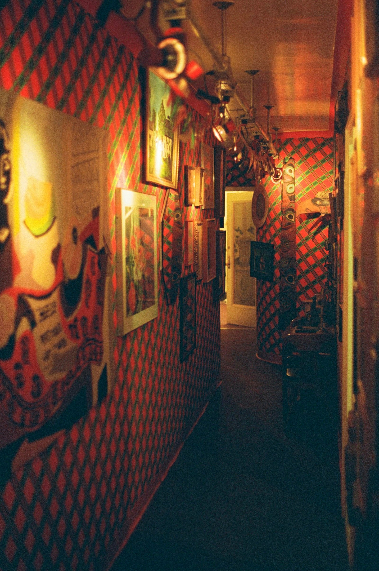 A narrow hallway wallpapered in a red and green diamond trellis pattern, lined with framed prints and posters under a row of hanging ceiling lights